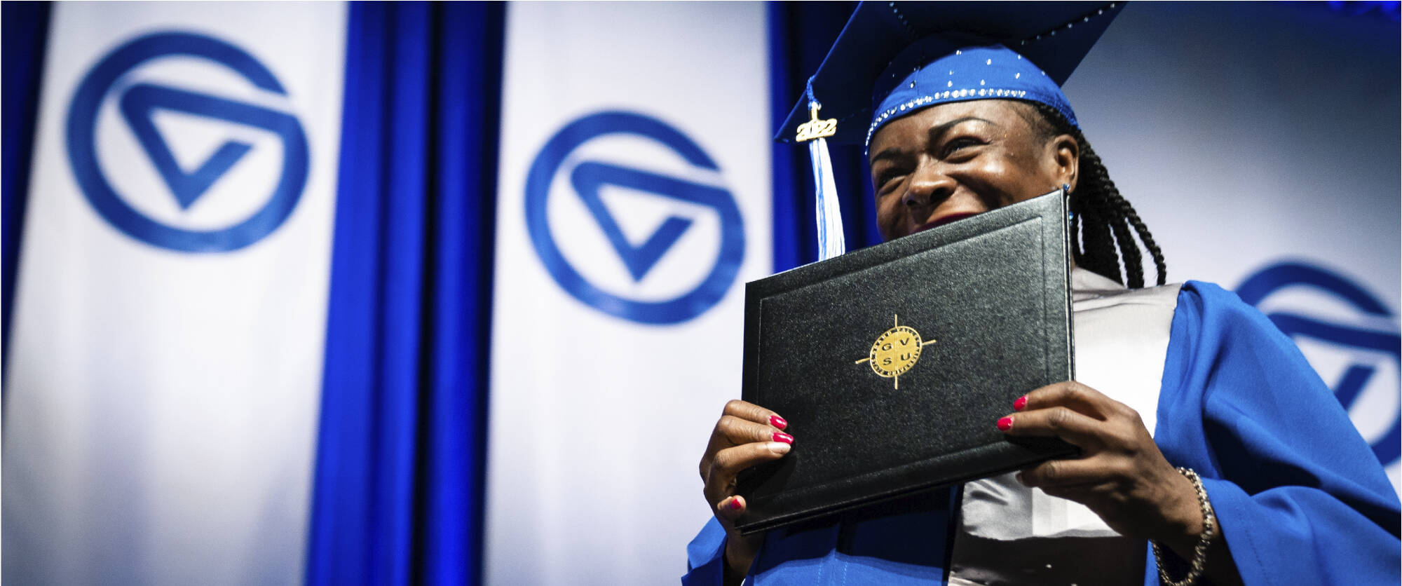 Smiling graduate holding their GVSU diploma at Commencement and wearing a cap and gown